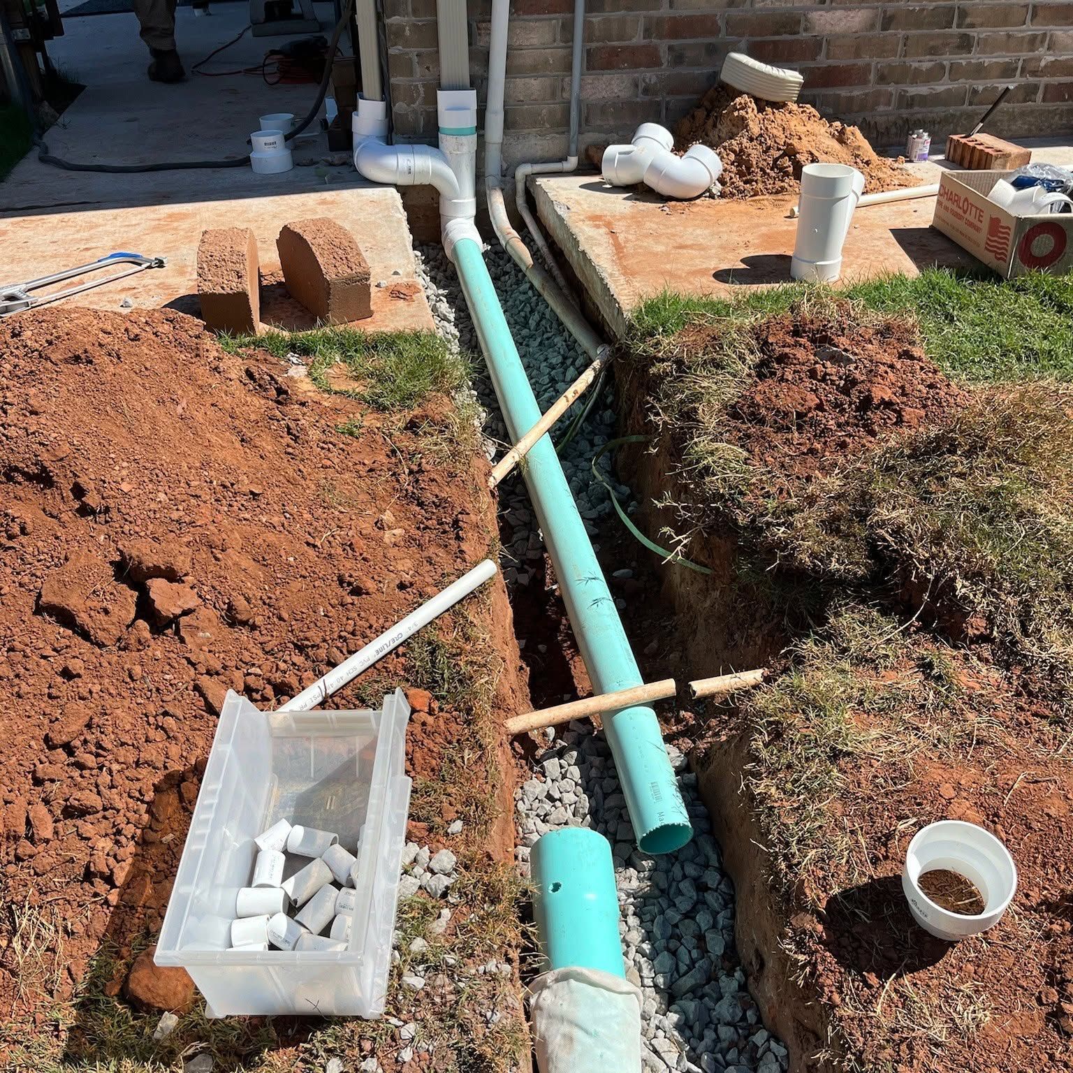 Open trench with blue PVC pipes and white fittings in red dirt at a construction site