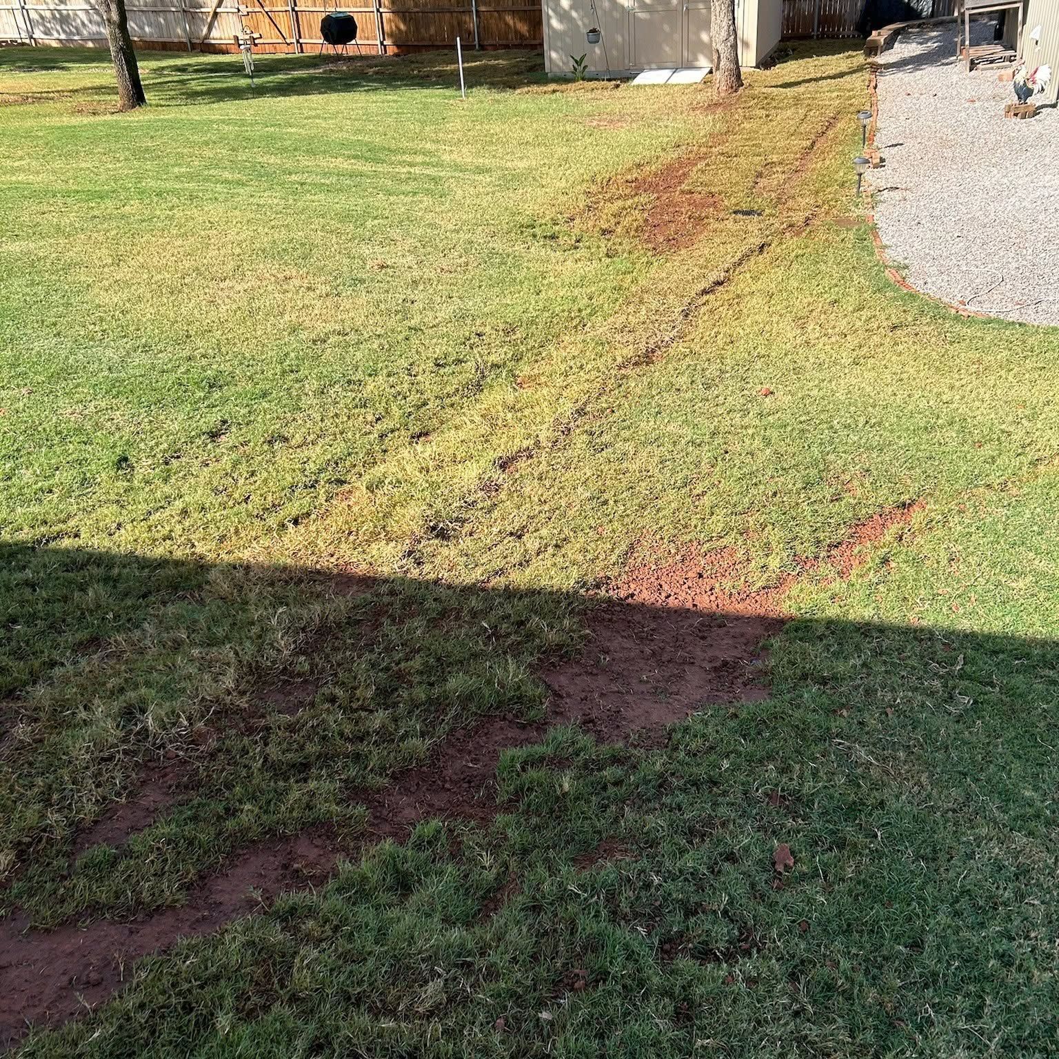 Grassy yard with a muddy tire track curving beside a gravel driveway and house shadows