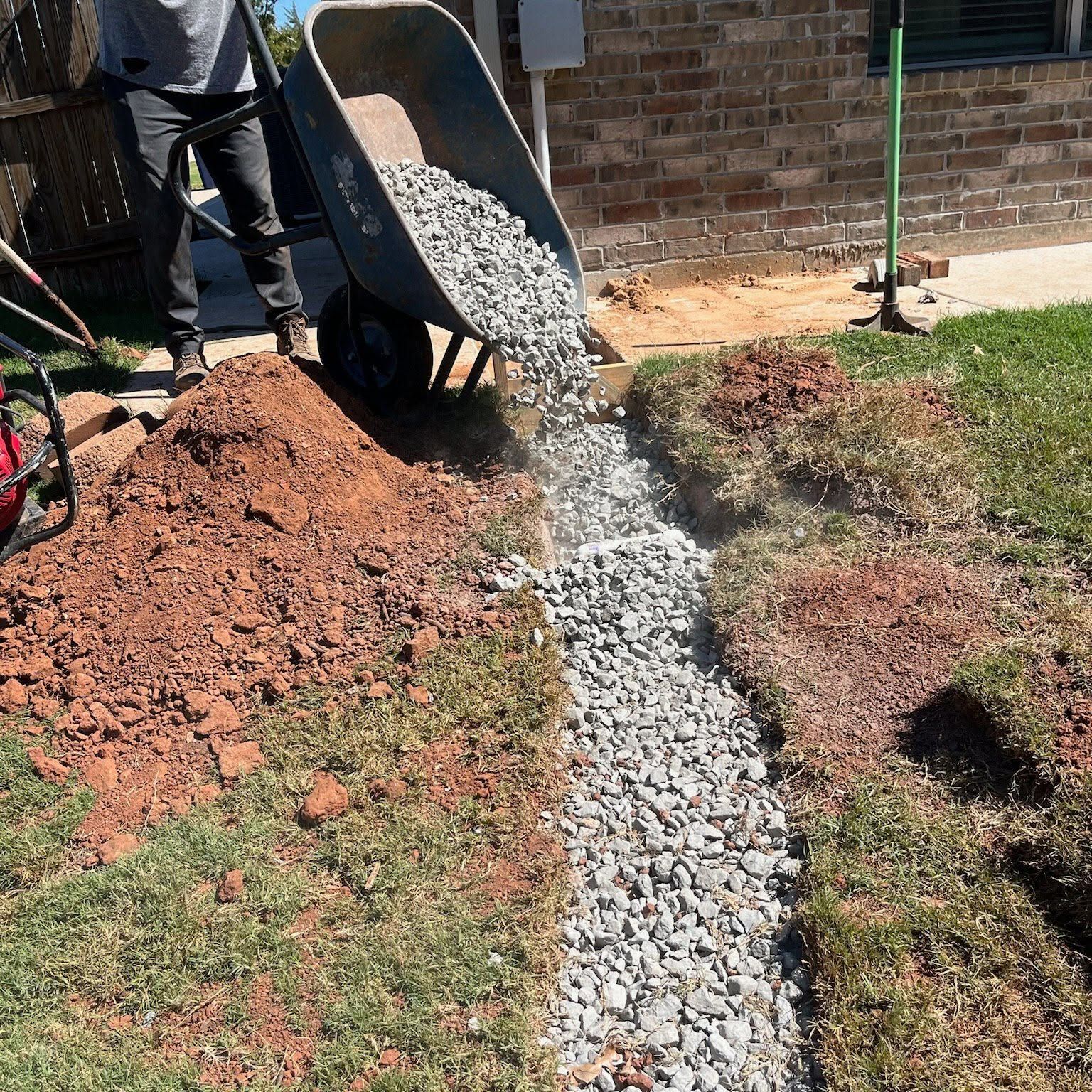 Person pouring gravel into a trench beside a brick wall, with a wheelbarrow and dirt piles in a yard.