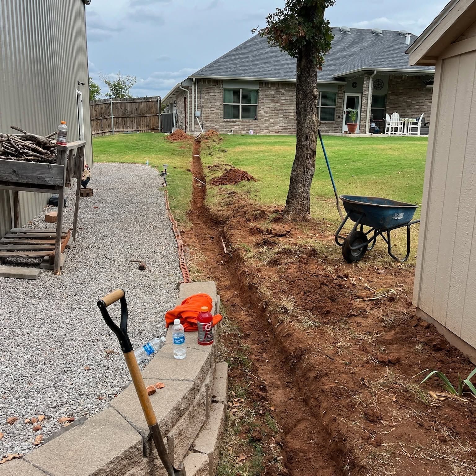 Backyard trench beside a house, with a shovel, wheelbarrow, and gravel path leading to a lawn.