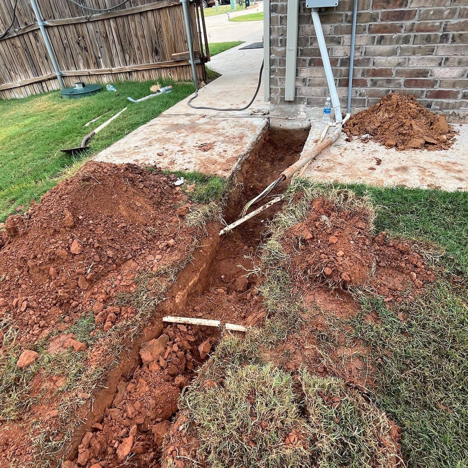 Backyard trench dug beside a brick house, with piles of red dirt and exposed pipes near a concrete patio