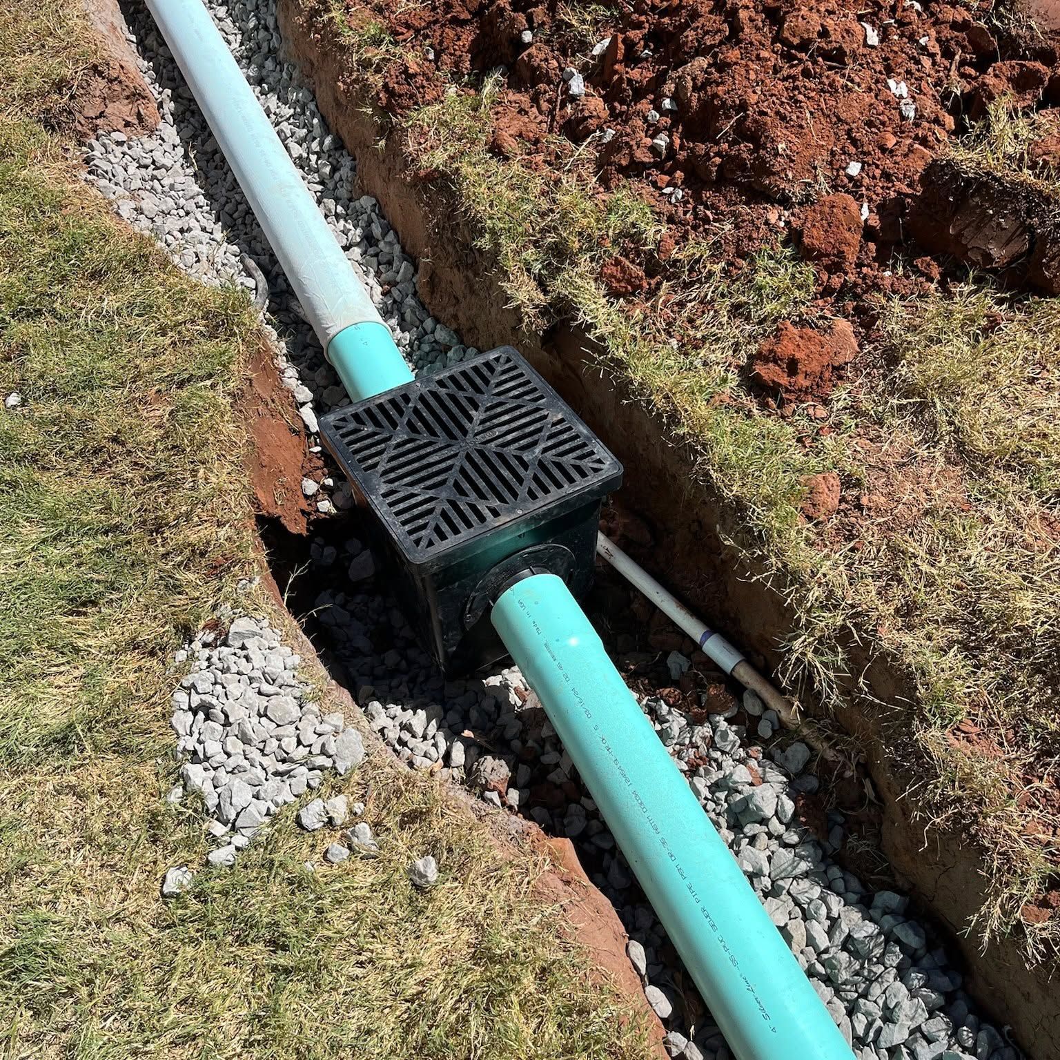 Black drainage pipe crossing a trench with a black grate, surrounded by soil and gravel