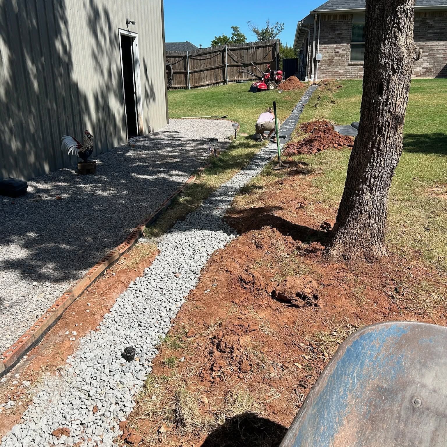 Backyard with a gravel path beside a shed, tree, and piled mulch under bright sunlight