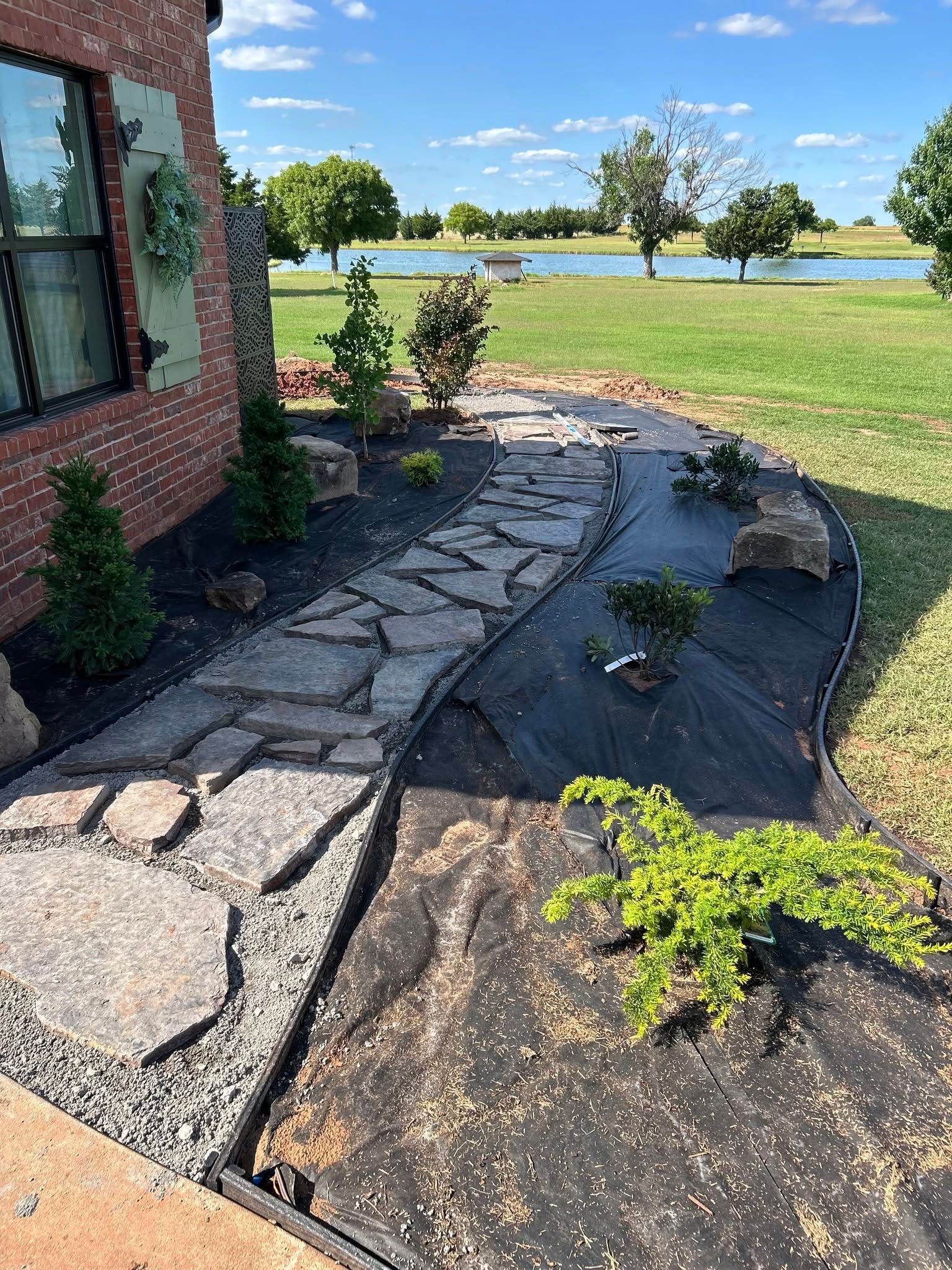 Stone garden path beside a brick house, with small plants and a lake view under a sunny blue sky