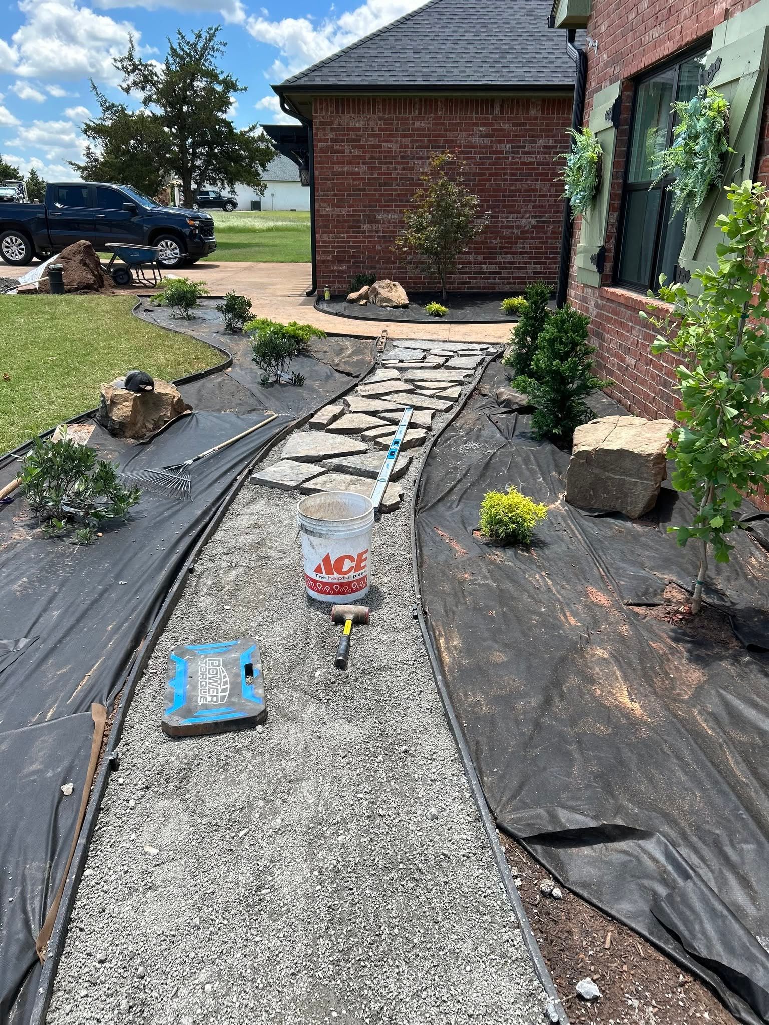 Gravel garden path under construction beside a brick house, with black landscape fabric and small plants.