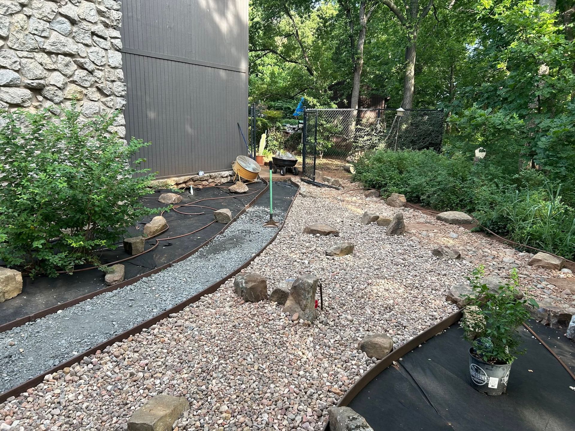 Gravel garden path beside a stone wall, bordered by shrubs and trees, with a potted plant on the right.