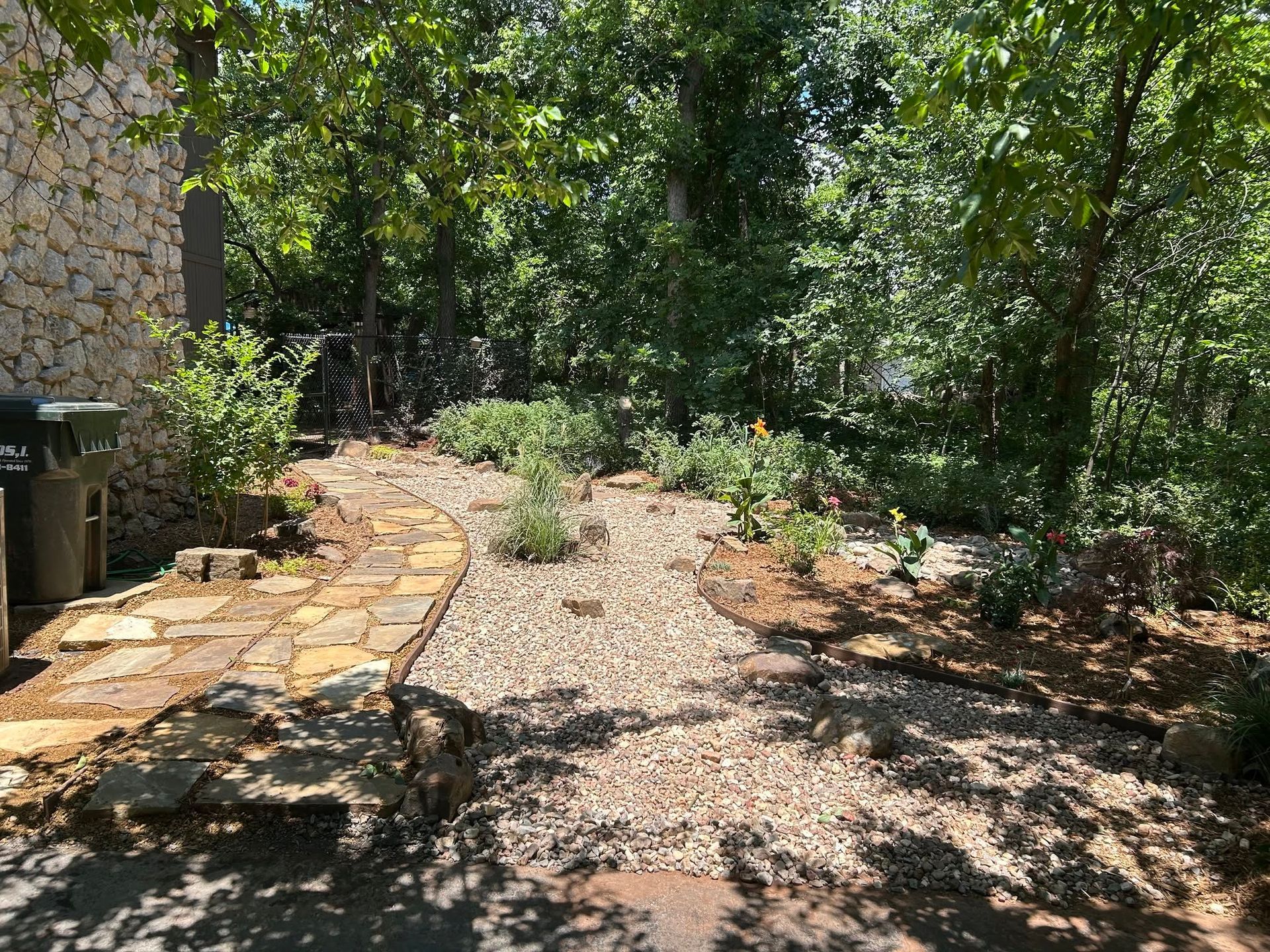 Stone patio beside a gravel garden path with trees, shrubs, and dappled sunlight.