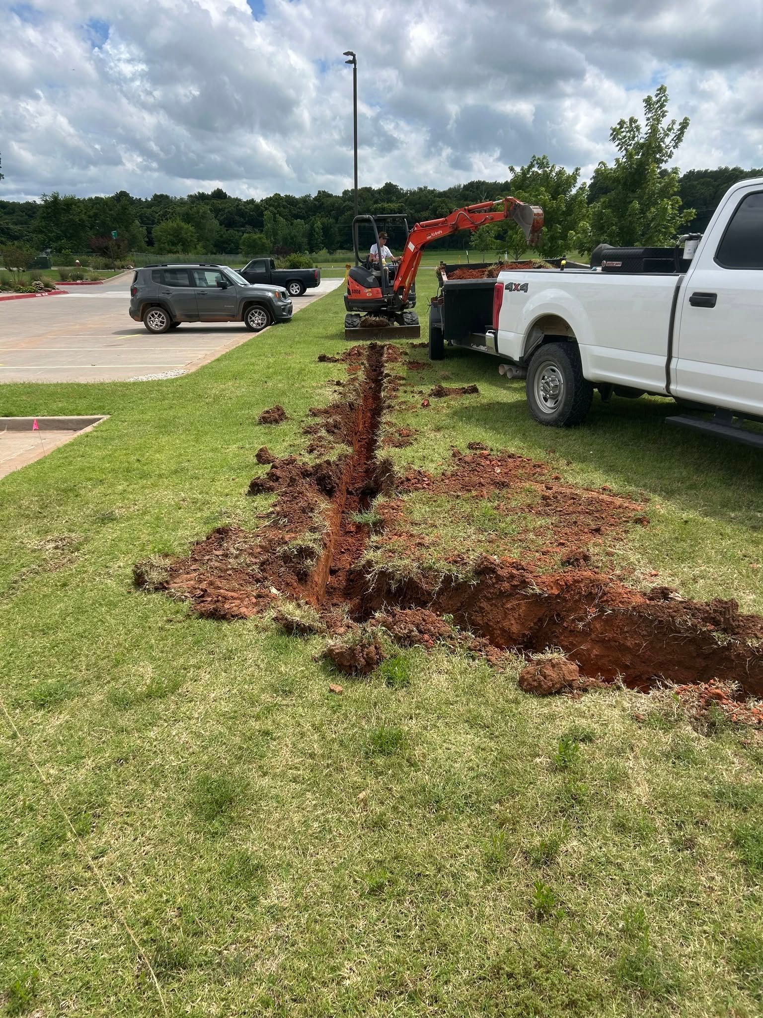 Trencher cutting a narrow trench in a grassy roadside beside parked trucks and a car.
