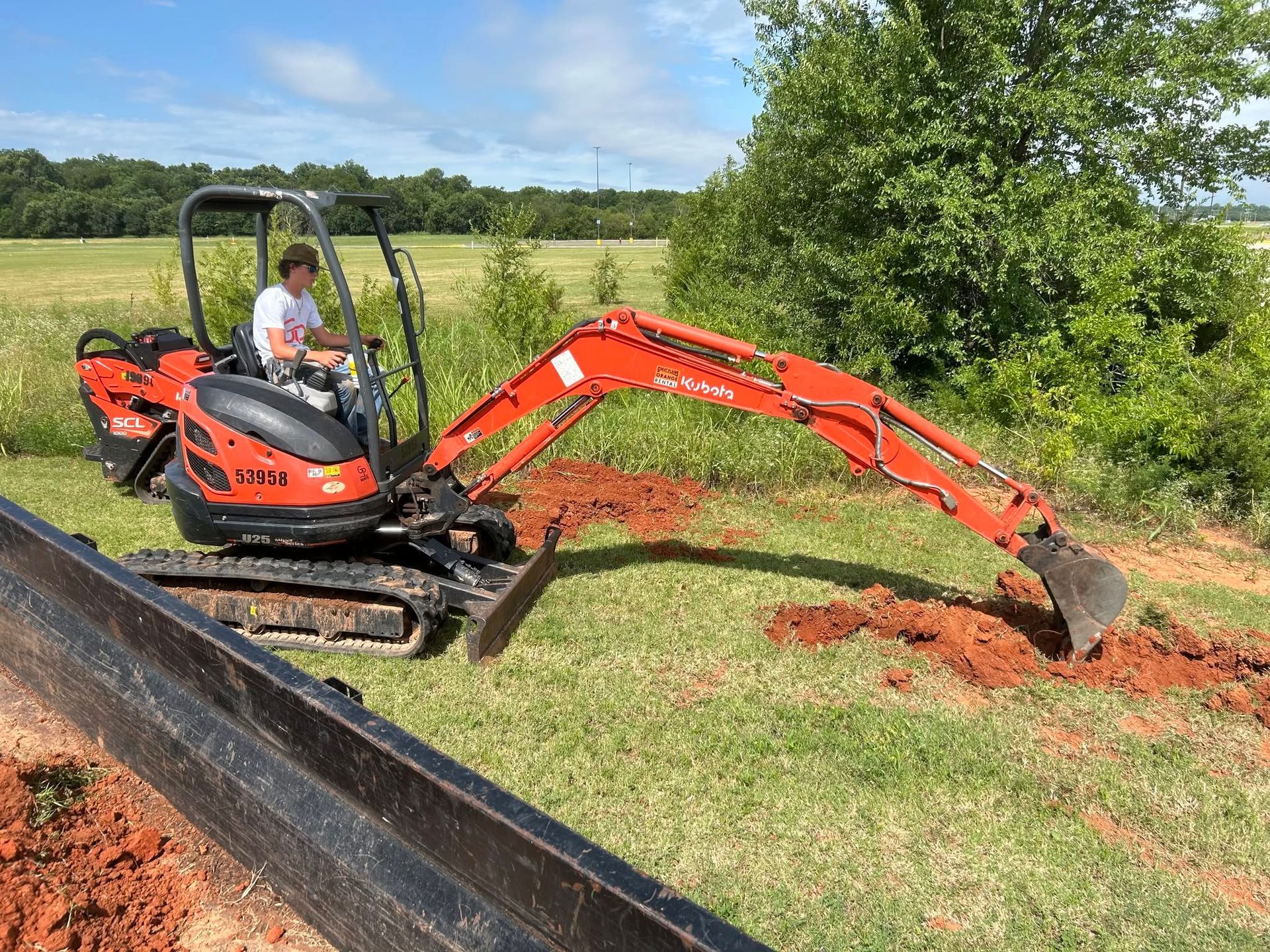 Small orange excavator digging a trench beside a black barrier in a grassy field.