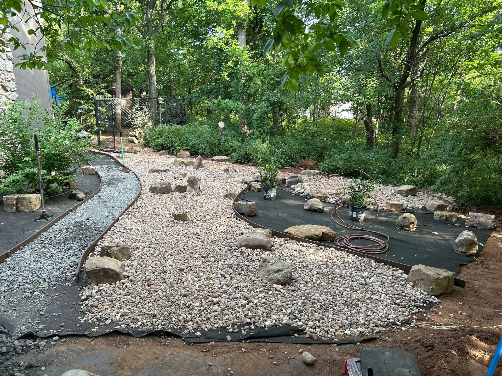 Garden path of gray gravel bordered by black edging, with rocks and green trees in the background