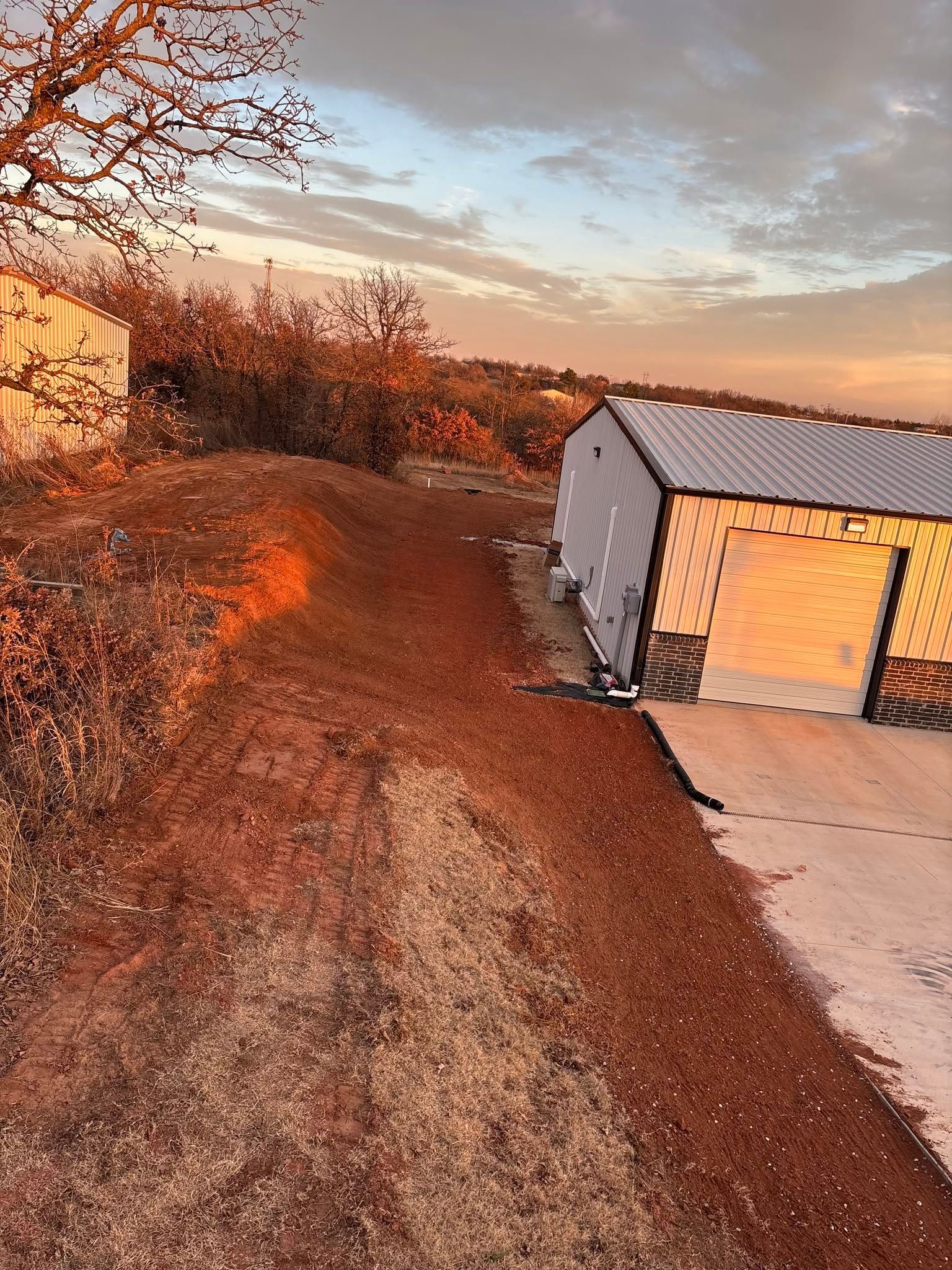 Rural dirt path beside a white shed at sunset, with trees and a concrete driveway.