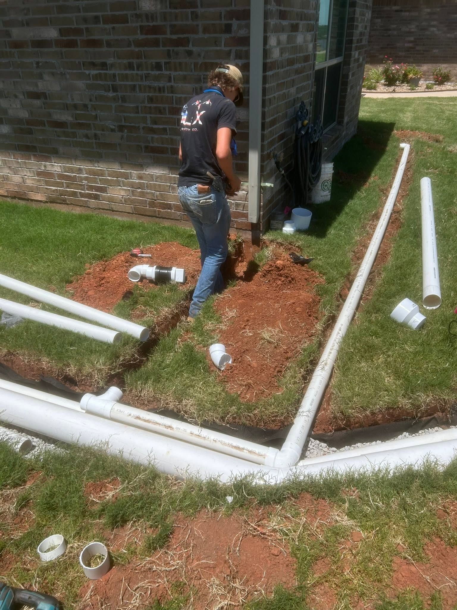 Person working on white PVC pipes beside a house, digging in a mulched yard.