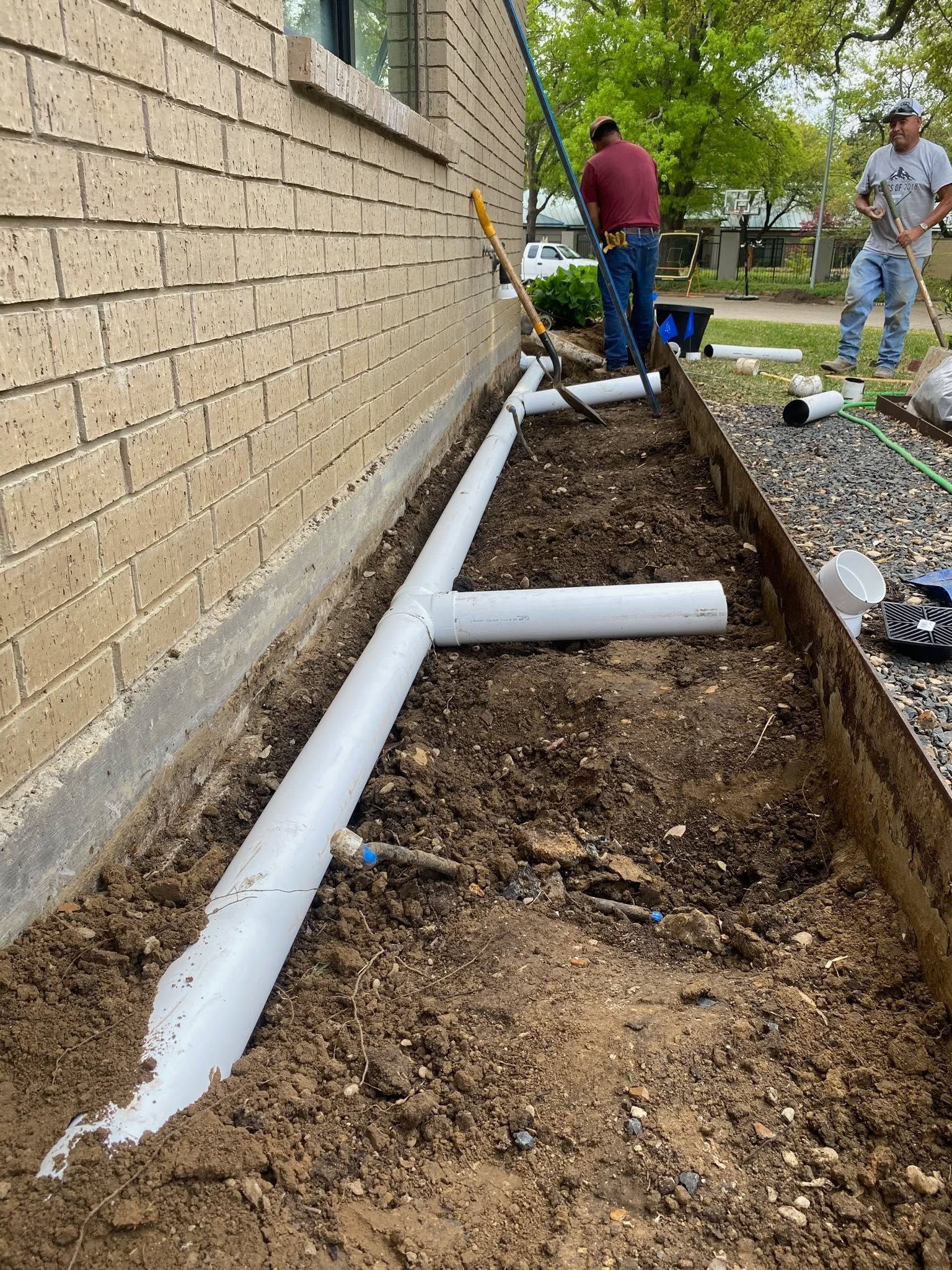 Workers installing white drainage pipes along a brick house foundation in a dirt trench