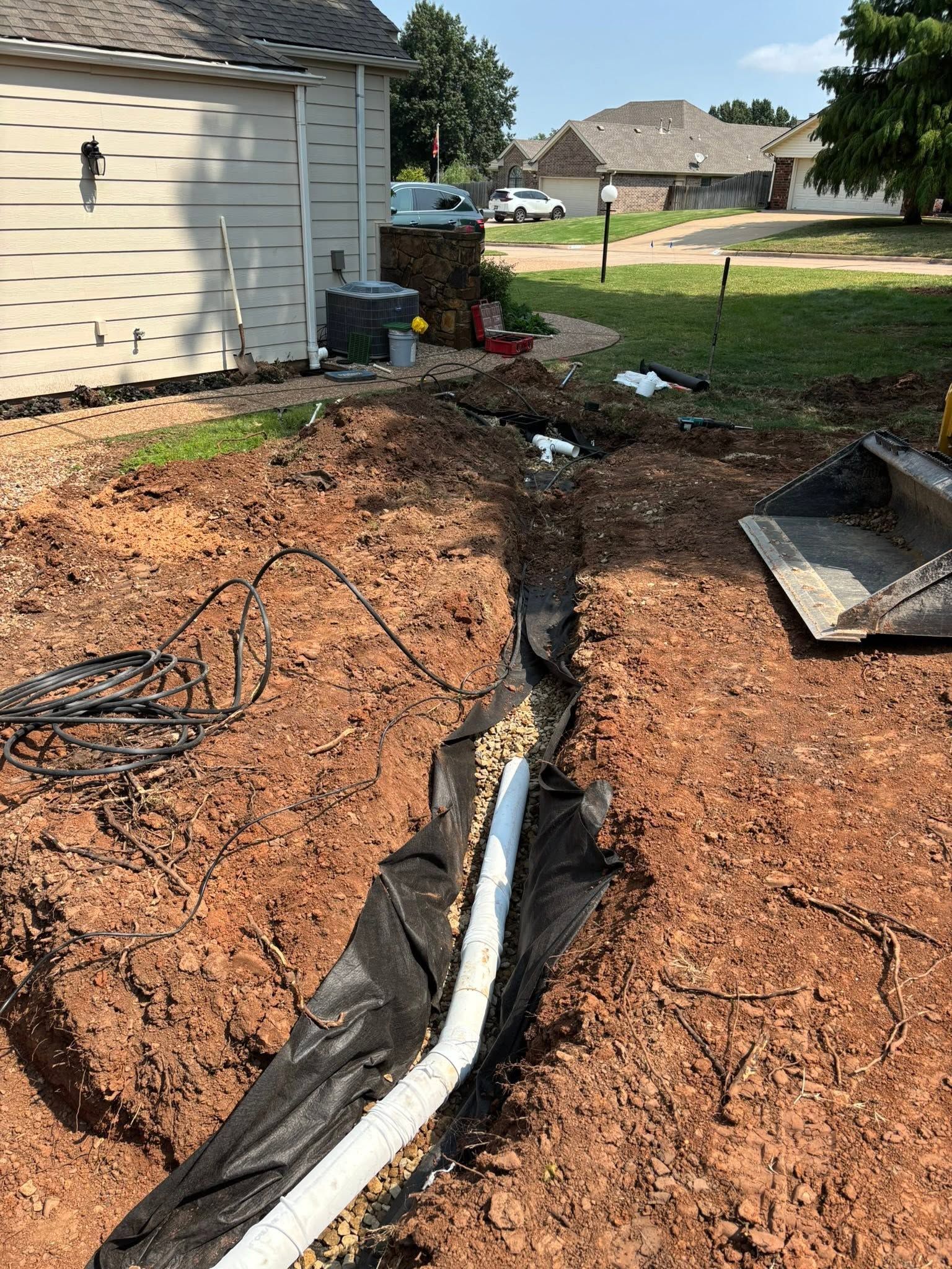 Backyard excavation trench with exposed white pipe beside a house and black landscape fabric