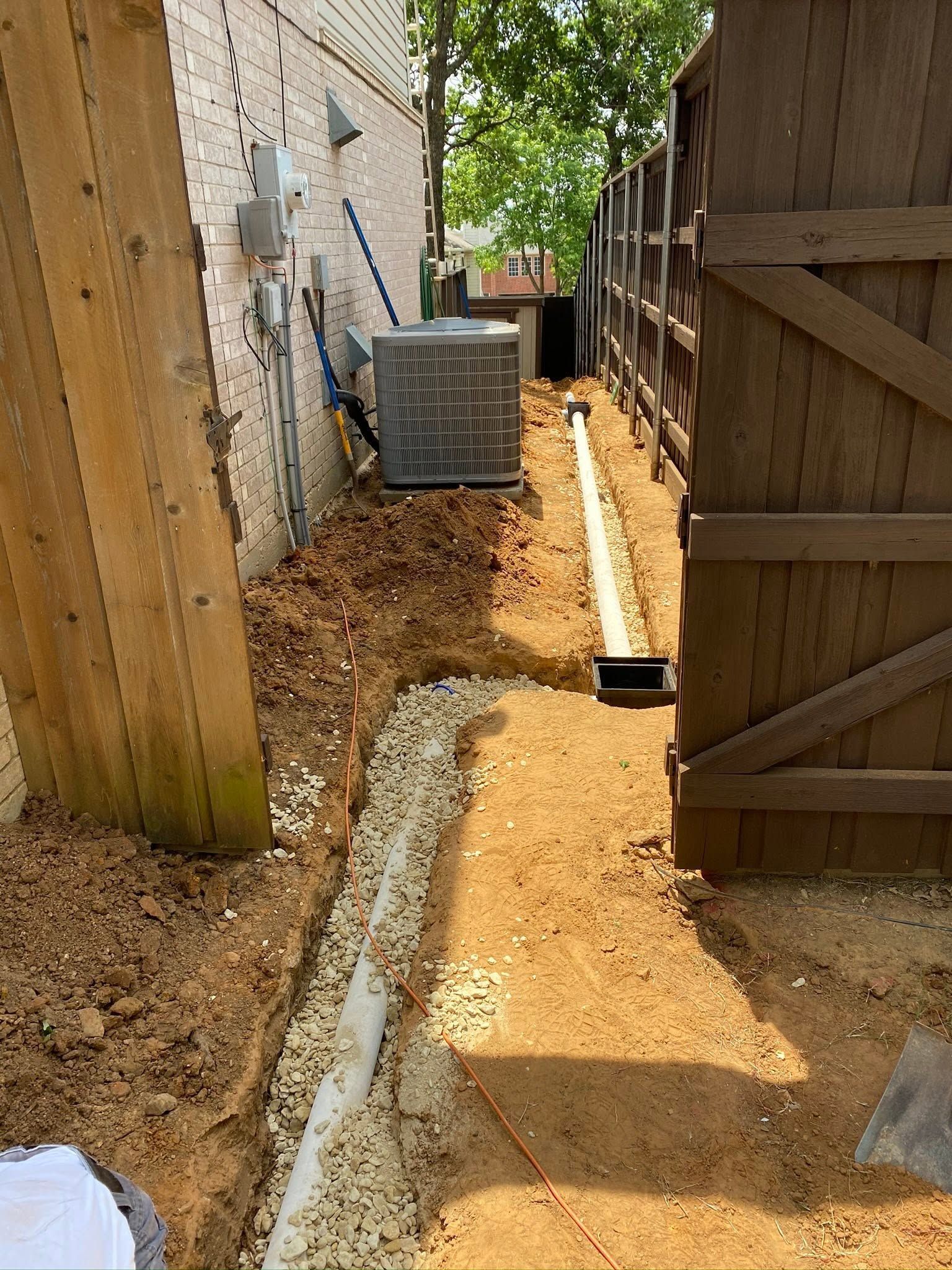 Narrow dirt walkway between fences beside a house, with a concrete drainage strip and a metal AC unit ahead