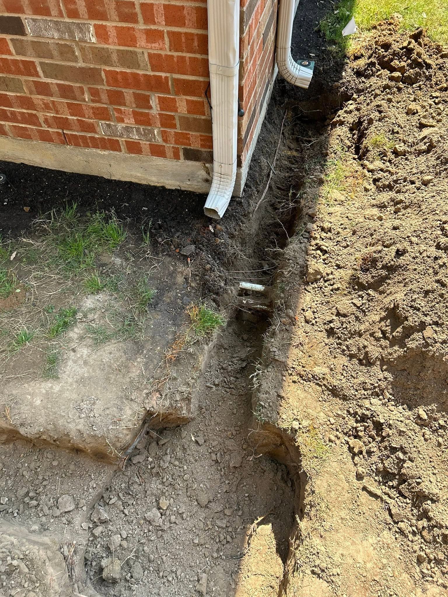 Brick building corner beside a narrow muddy trench or drainage channel along a gravel path