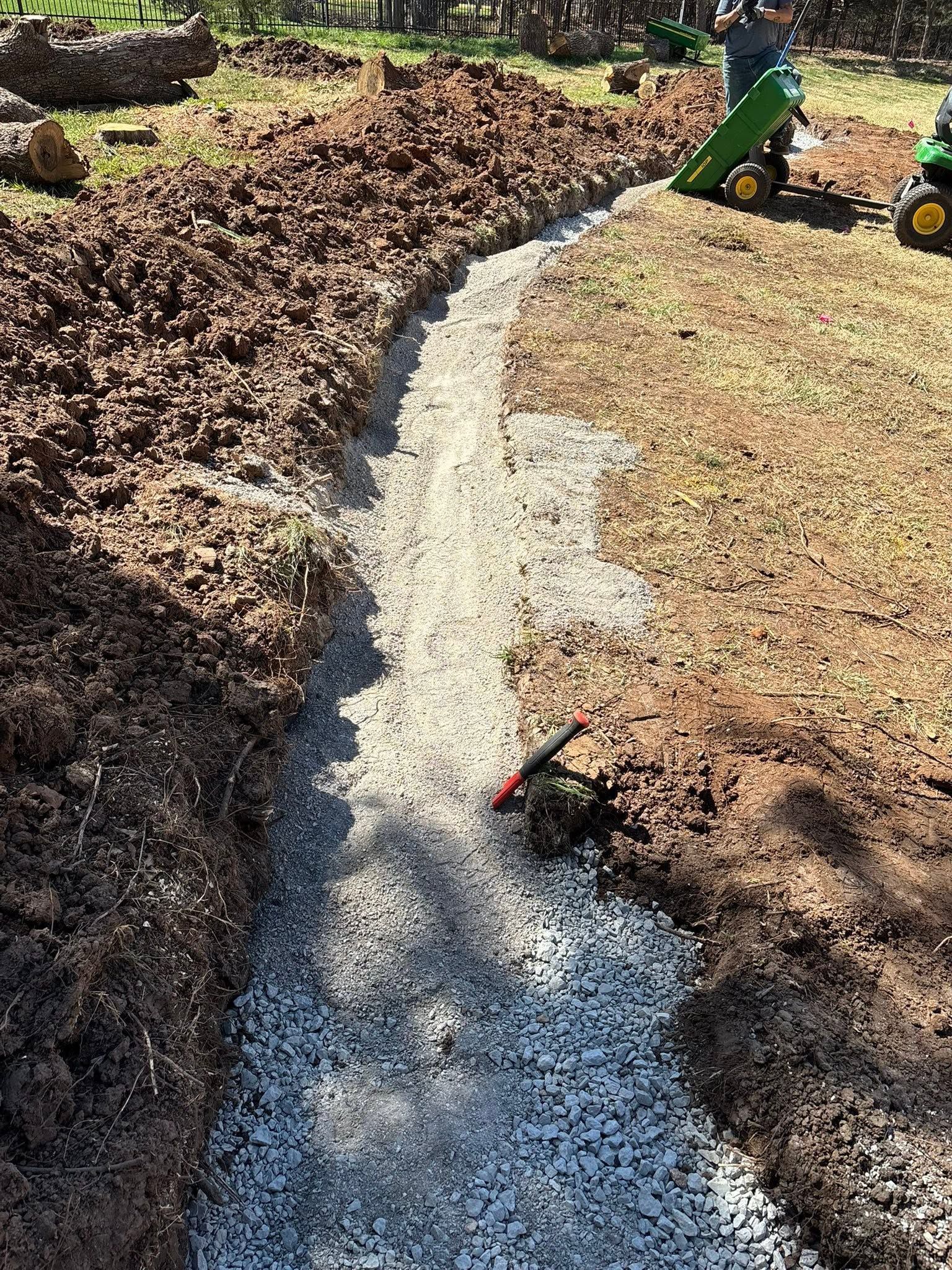 Fresh gravel path being installed through a grassy yard beside dark mulch beds, with a small tractor nearby