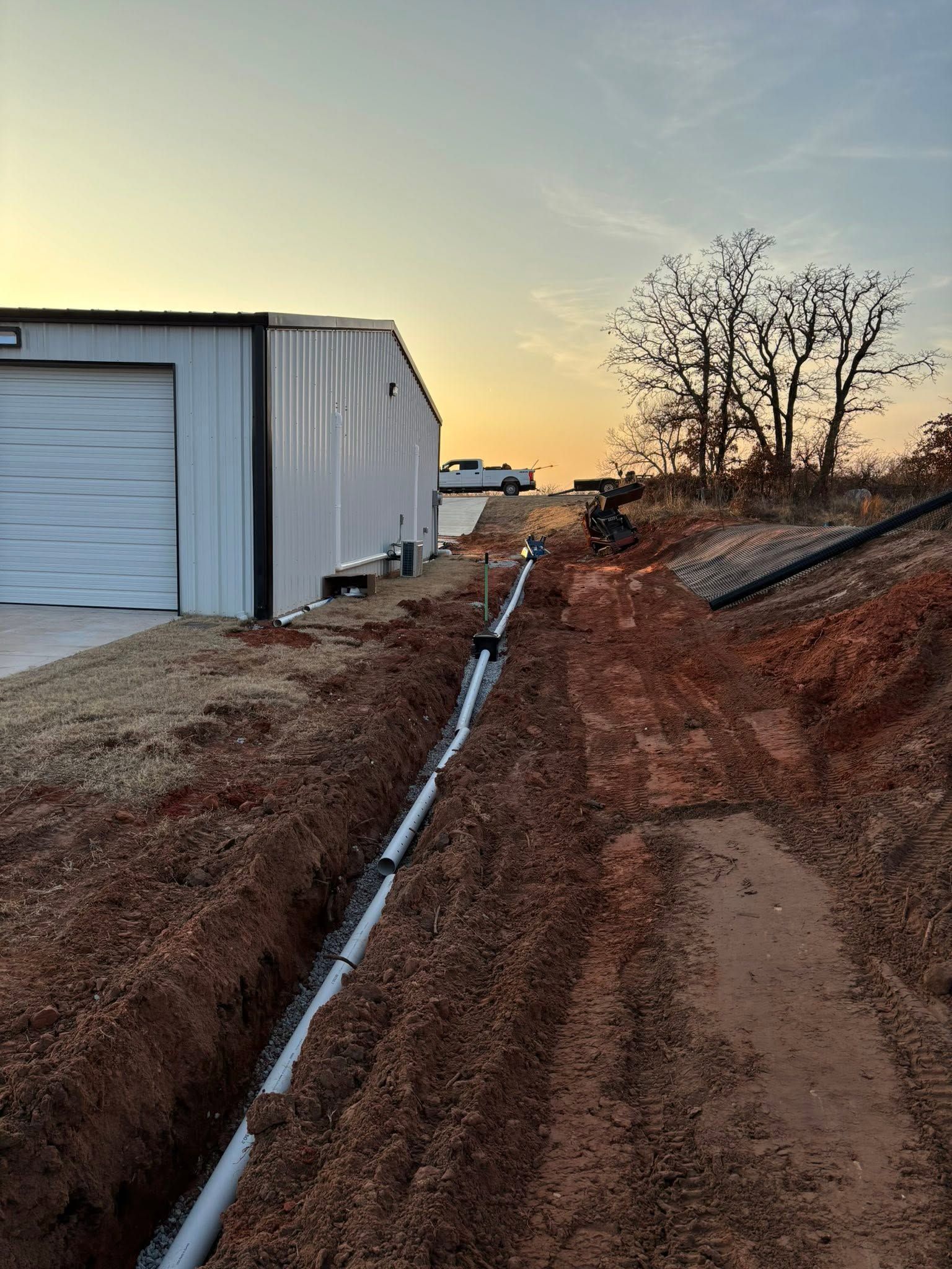 Dirt path beside white warehouse at sunset, with drainage ditch and bare trees on the right.