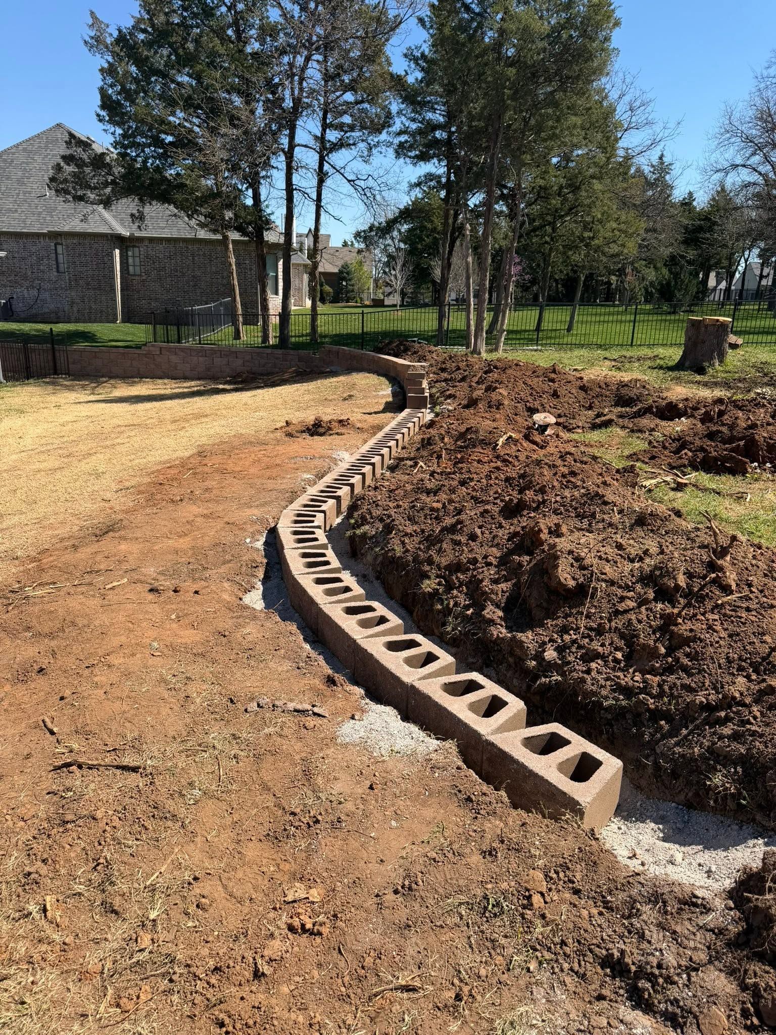 Curved cinder block edging beside a dirt path and trench in a wooded yard under construction