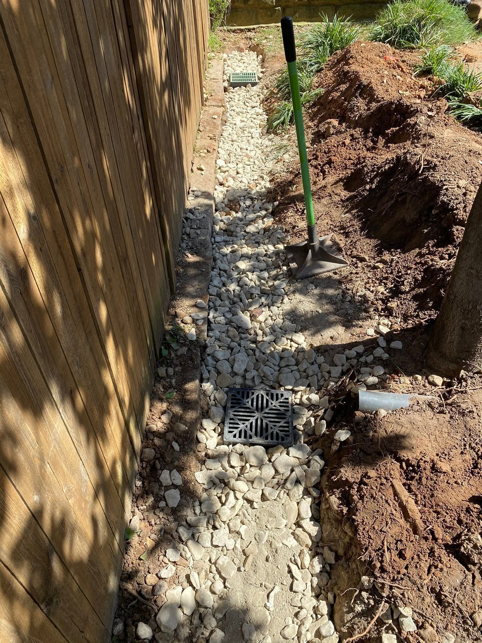Narrow gravel trench beside a fence, with pipes and a small drainage grate between dirt mounds.