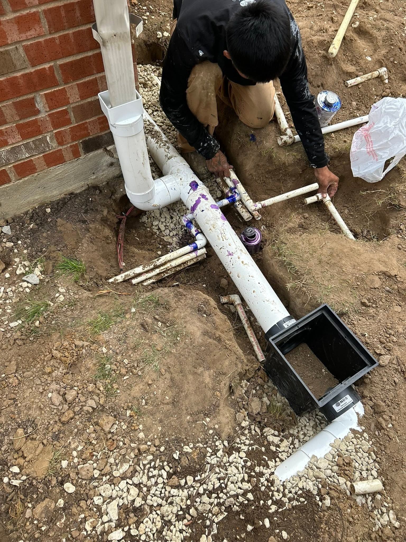 Worker kneeling by a white PVC pipe and black drain box, installing plumbing near a brick wall outside.