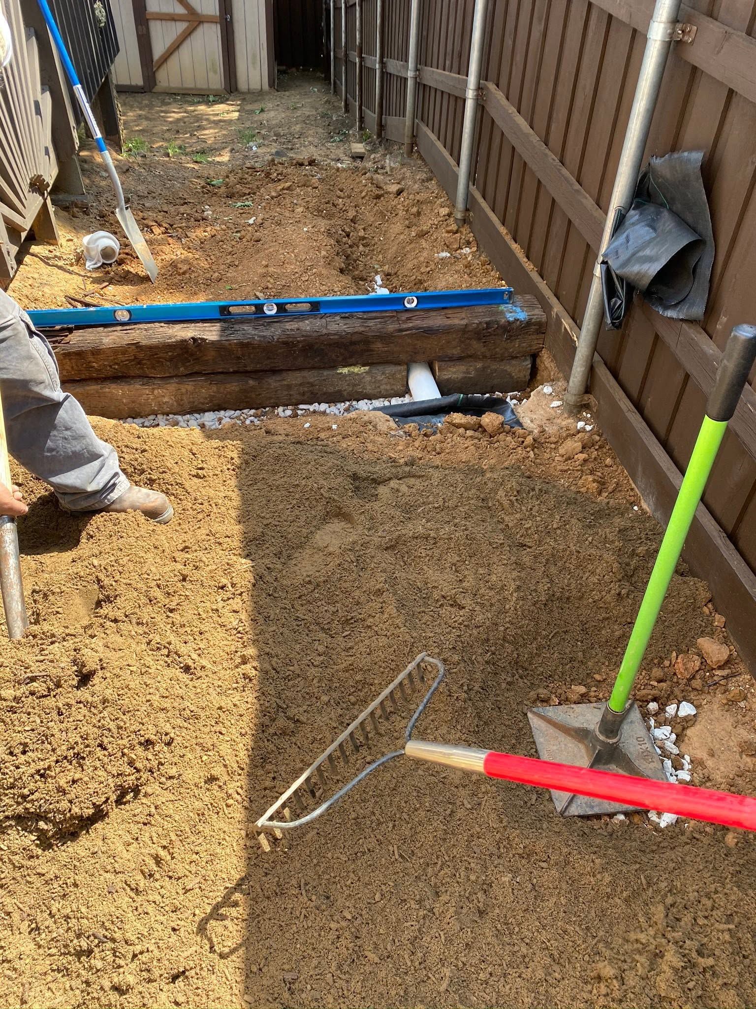 Narrow construction trench with gravel, fencing, and blue, green, and red tools laid across it.