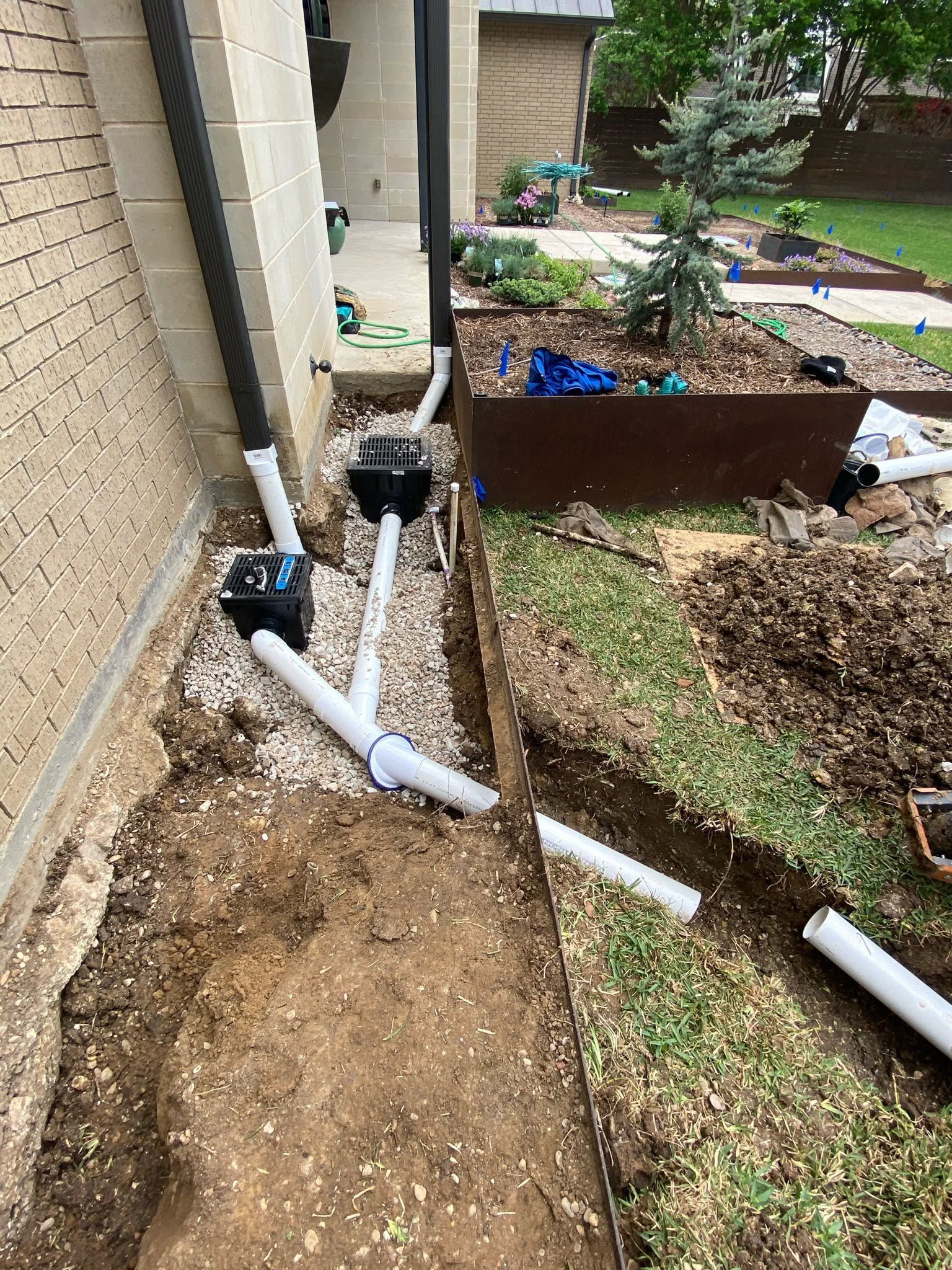 Damaged white drainage pipes along a house foundation beside a dirt trench and garden bed