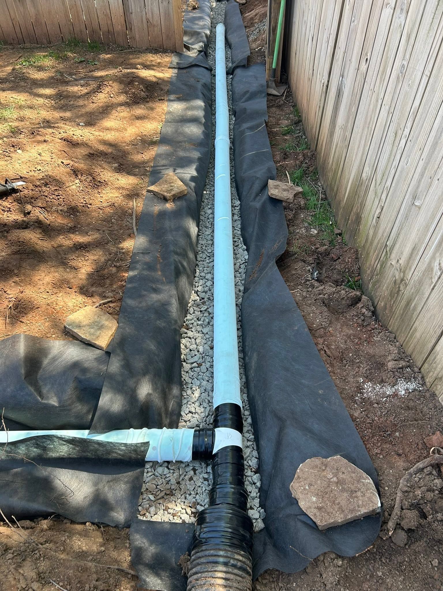 Drainage pipe running between black landscape fabric along a narrow dirt strip beside a fence