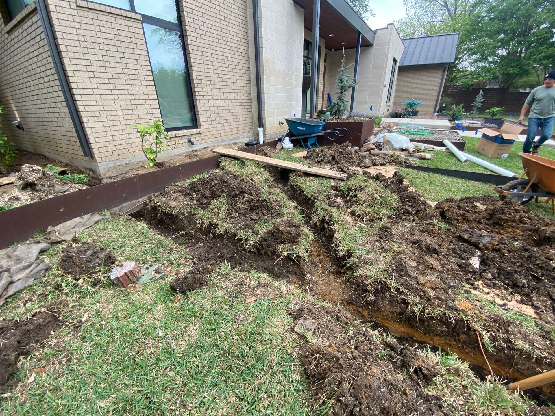 Severe soil erosion beside a brick house, with exposed trenches and scattered debris in the yard.