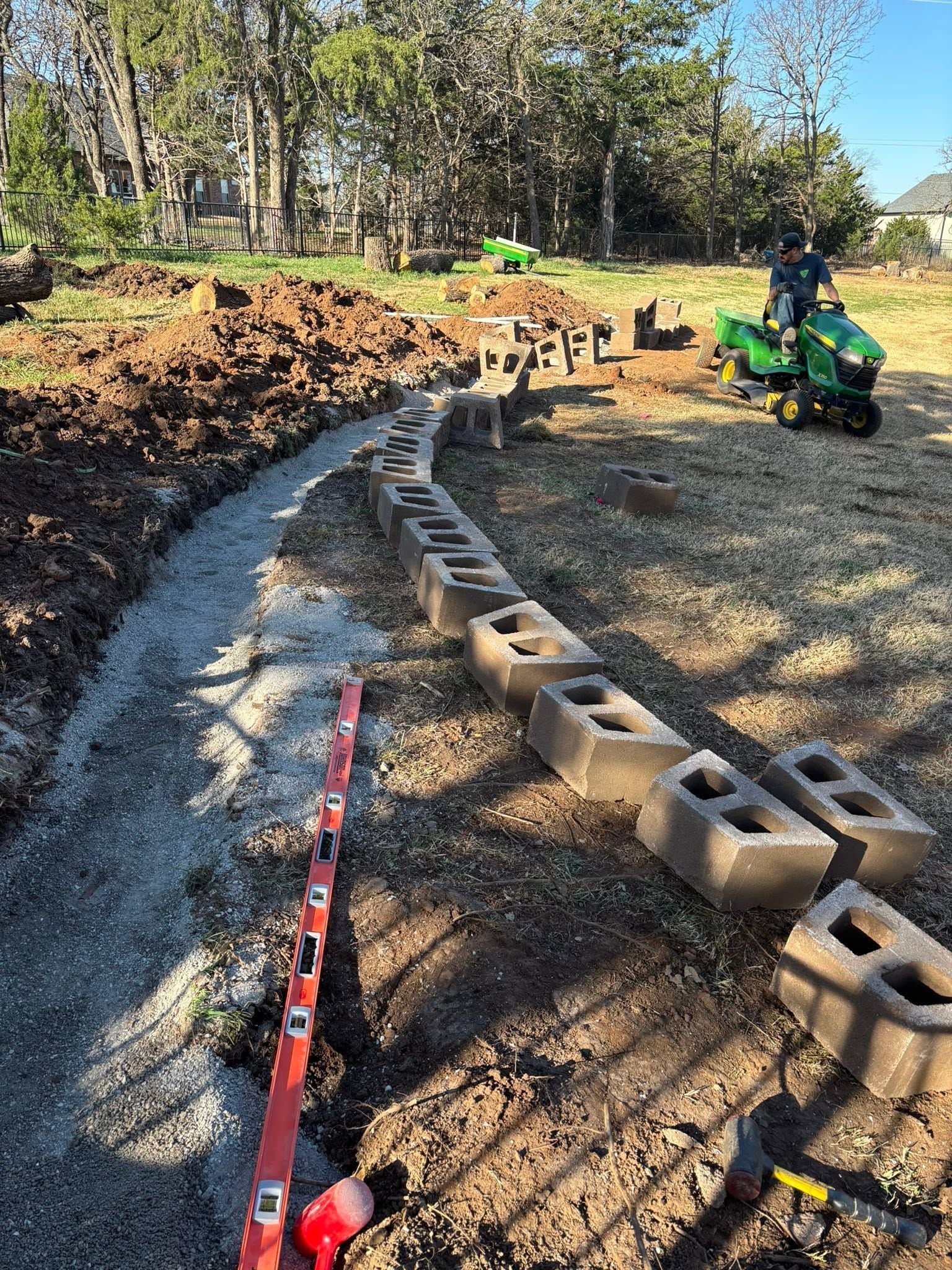 Curved cinder-block path with gravel in a wooded yard, with tools and a green riding mower nearby