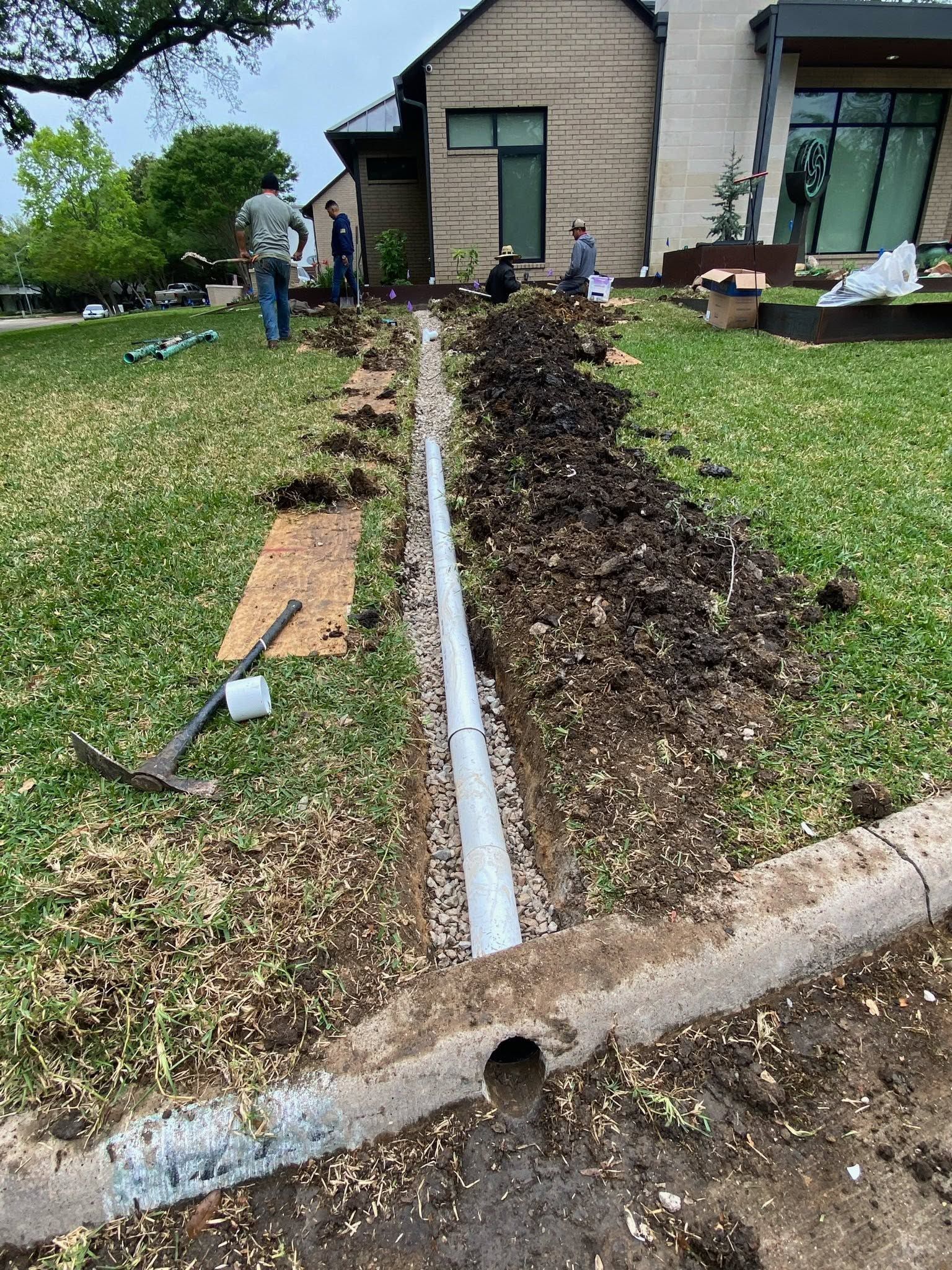 Broken sidewalk and exposed trench beside a house, with workers and debris in a front yard.