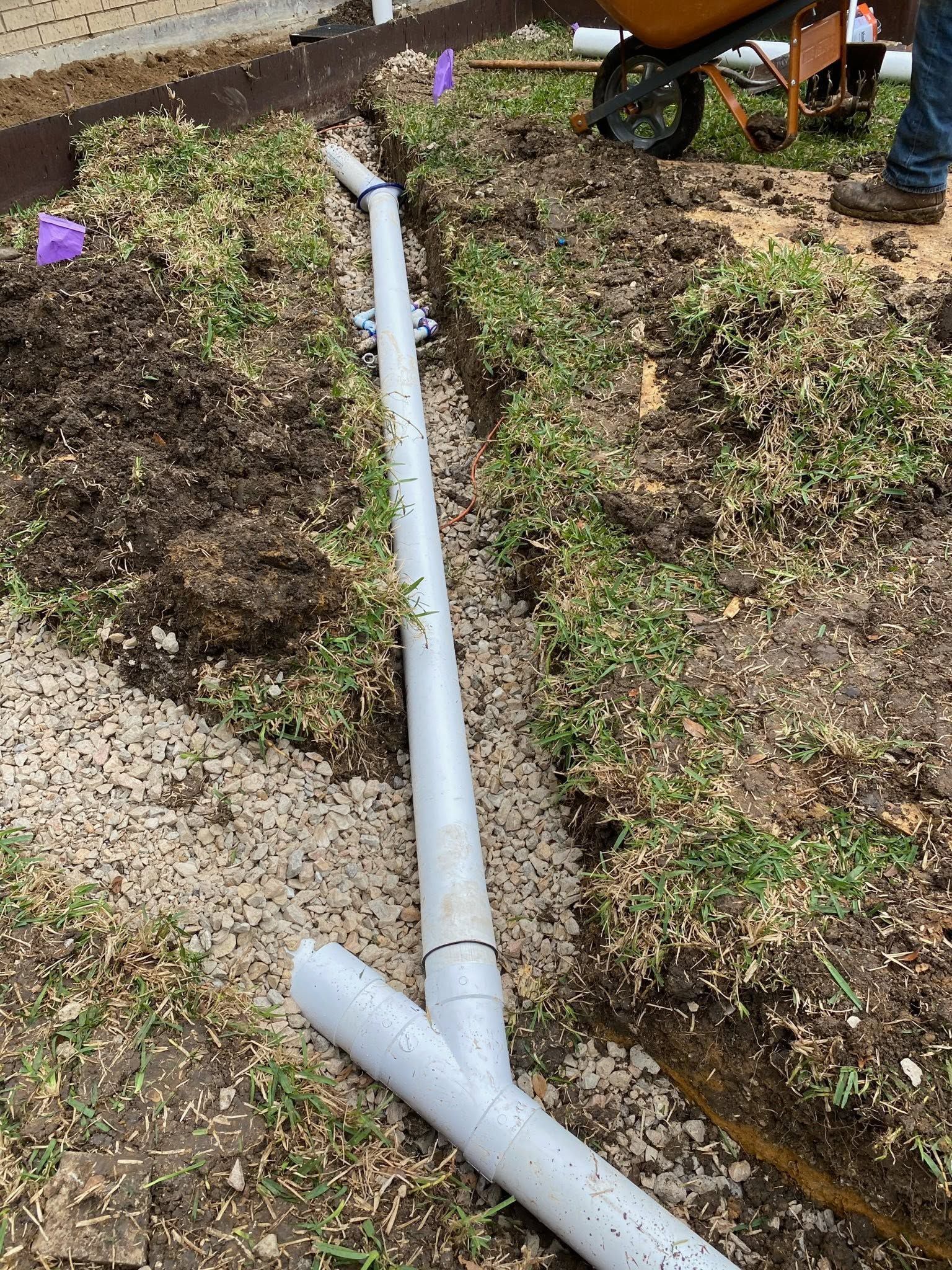 White drainage pipe runs down a dirt slope beside gravel and grass, with a wheelbarrow at the top.
