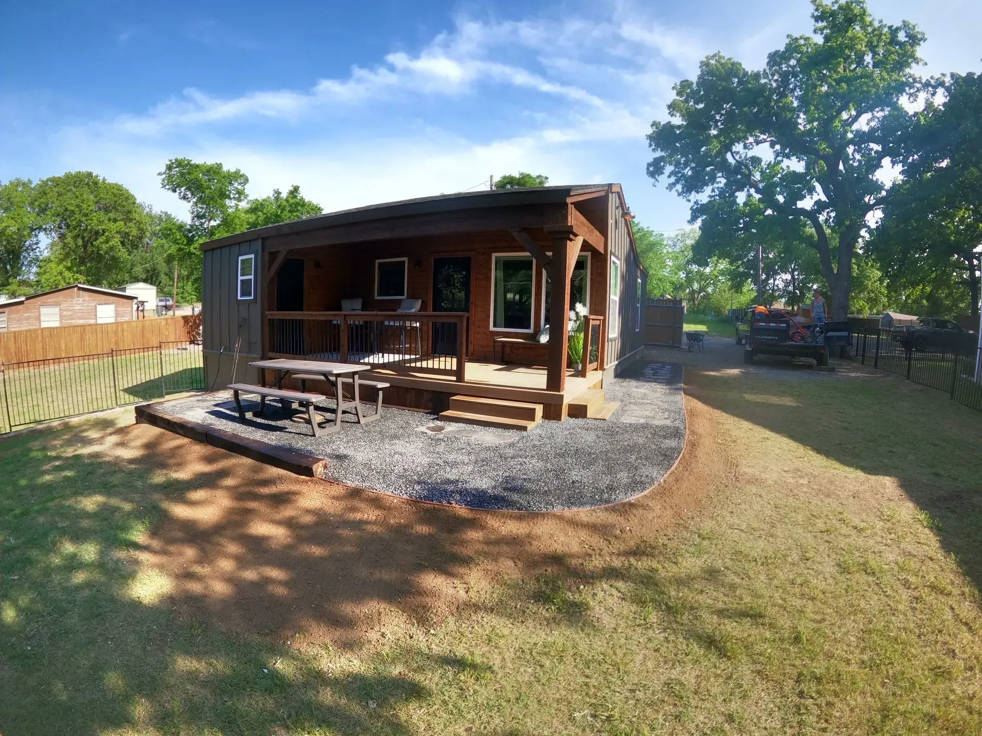 Wooden cabin with a front porch and gravel patio in a sunny backyard, surrounded by trees and lawn.