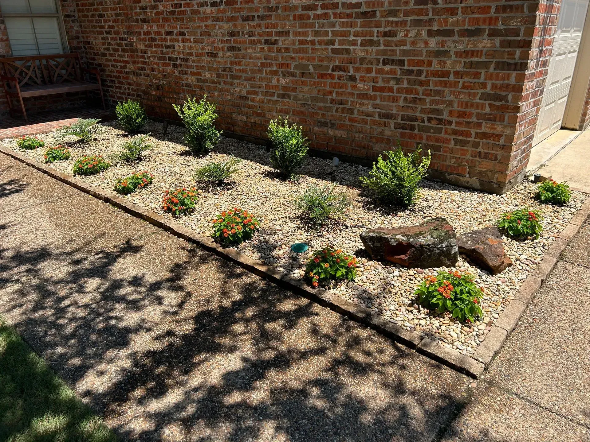 Gravel landscaping bed with small shrubs and flowers beside a brick wall in sunlight