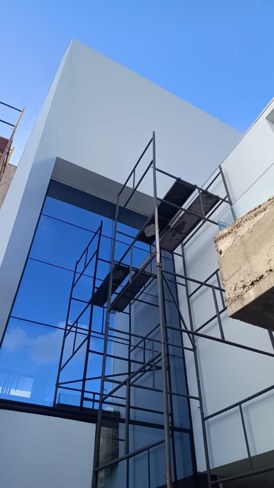 Fachada de edifício moderno com janelas azuis refletoras e andaimes de construção pretos contra um céu claro.