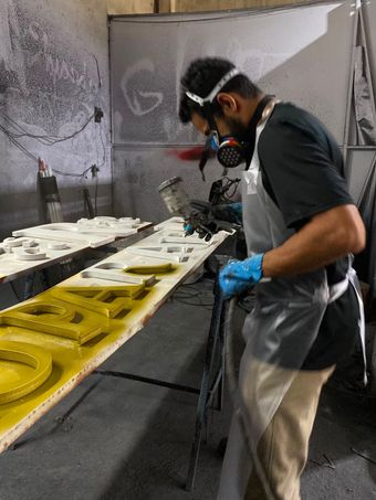 Operário em uma cabine de pintura aplicando tinta em painéis amarelos e brancos, usando máscara respiratória e luvas.
