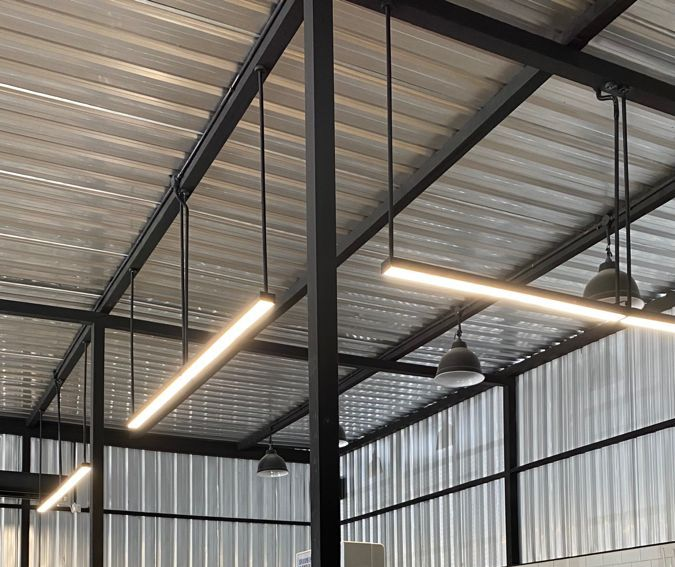 Teto industrial com vigas metálicas expostas, cobertura ondulada e luminárias pendentes no interior de um armazém.