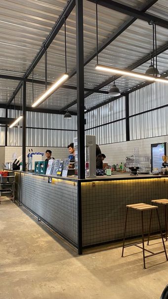 Balcão de café industrial com grade de malha metálica, luzes pendentes e banquetas em um espaço estilo armazém.
