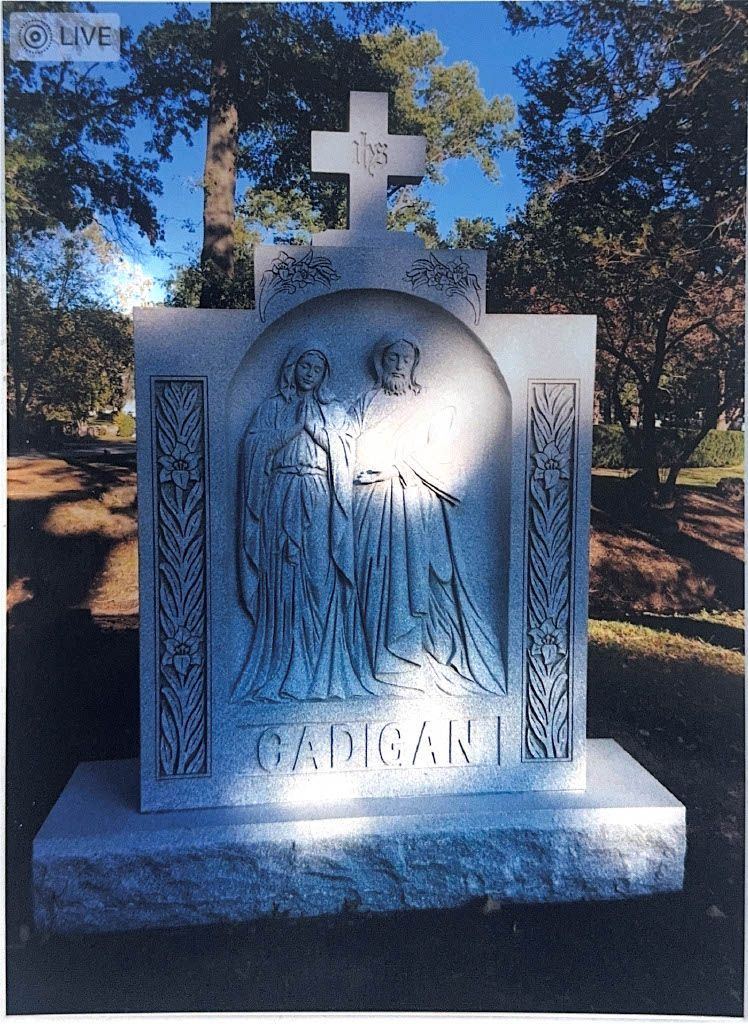 White stone memorial with a cross and engraved figure, standing in a wooded cemetery.