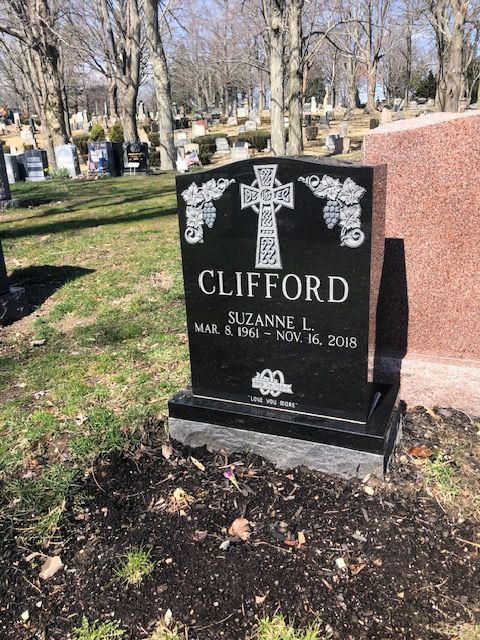 Black granite gravestone with cross and floral carvings in a cemetery, engraved “Clifford” and “Suzanne L.”