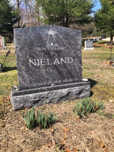 Granite grave marker reading “NIELAND” in a cemetery, with flowers and pine trees nearby