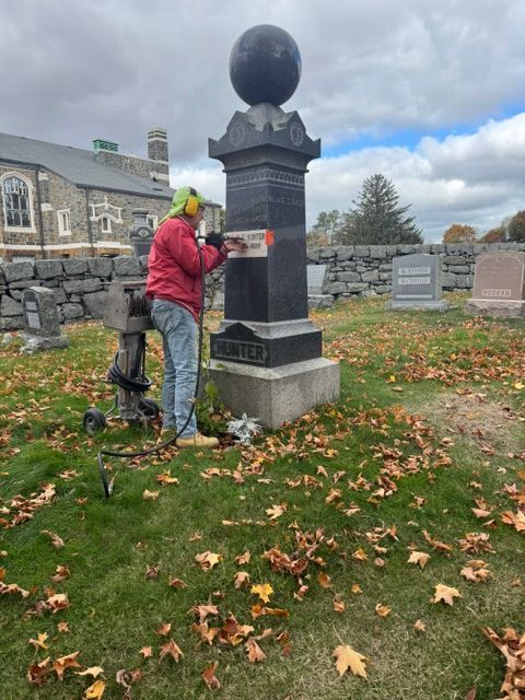 Worker cleaning a black monument in a cemetery with fallen autumn leaves.