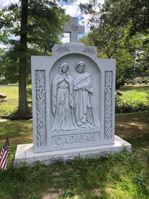Stone gravestone with relief of two figures and the name “CADGAN” in a grassy cemetery