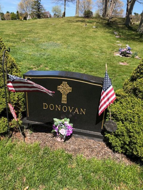 Black memorial headstone for Donovan with flowers and two small American flags on a grassy hillside