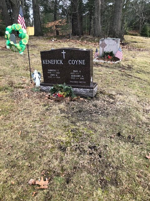 Gravestone in a grassy cemetery with wreaths, flowers, and an American flag nearby.
