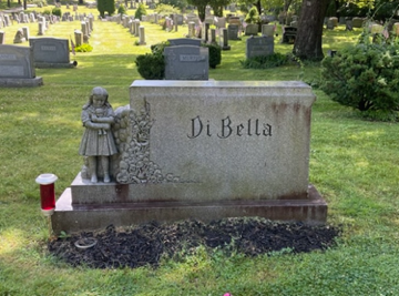Gravestone marked “Di Bella” with a small child statue beside it in a cemetery.