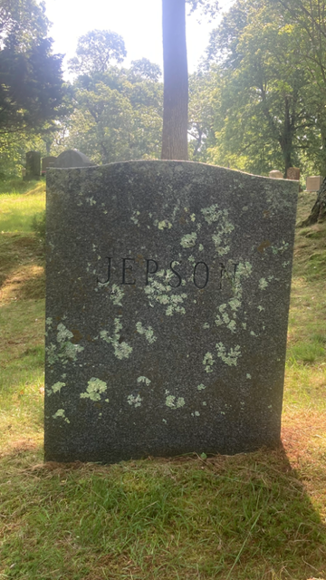Weathered dark stone memorial in a grassy cemetery, with a cross-shaped marker behind it.