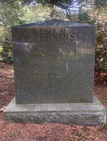 Weathered dark stone memorial in a grassy cemetery, with a cross-shaped marker behind it.