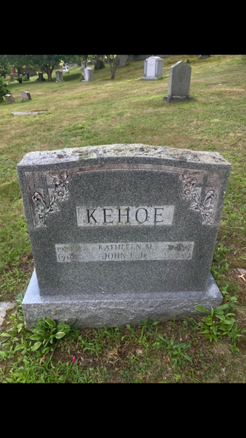 Weathered gray gravestone in a grassy cemetery, with other headstones in the background.