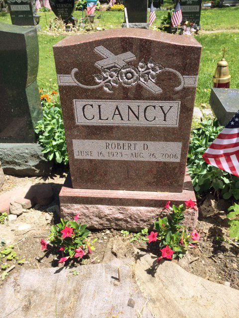 Clancy gravestone with flowers at a cemetery, featuring a cross and American flag nearby